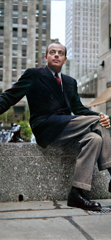 Antoine de Saint-Exupéry sitting on a stone ledge in a city, holding a cigarette.