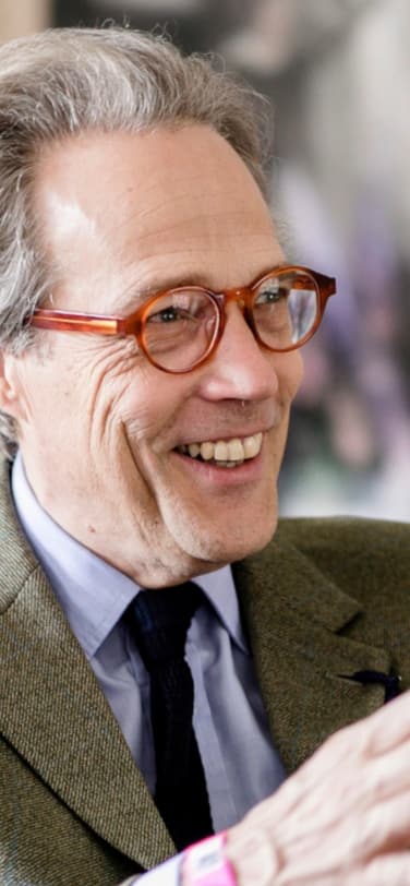 The 11th Duke of Richmond, wearing red glasses and a green jacket, seated and smiling while talking at an event.
