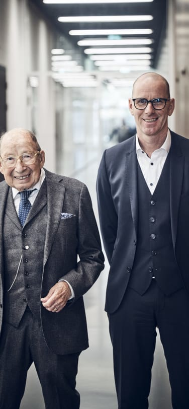 Two watchmakers in suits at a manufacturing center smiling at the camera