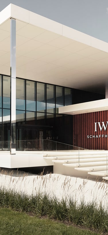 Das Manufakturzentrum ist ein modernes, zweistöckiges Gebäude mit einem flachen weißen Dach und großen Glasfenstern, das eine strukturierte Wand mit dem „IWC Schaffhausen“-Logo aufweist.