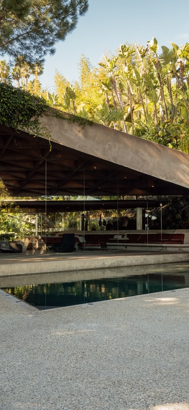 An exterior view of the Goldstein Residence reveals its unique modernist architecture, featuring an angular concrete roof, glass walls, and a swimming pool nestled within lush tropical landscaping.