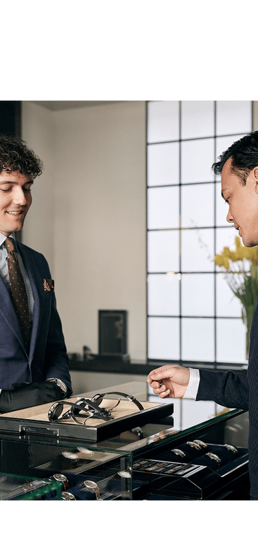A man shops for luxury watches at a boutique, assisted by a well-dressed salesman behind a glass display case.