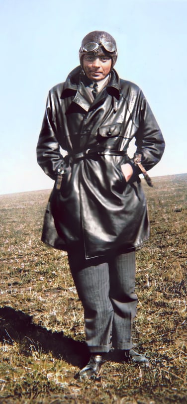 Antoine de Saint-Exupéry in a leather aviator coat, helmet, and goggles, standing in a grassy field.
