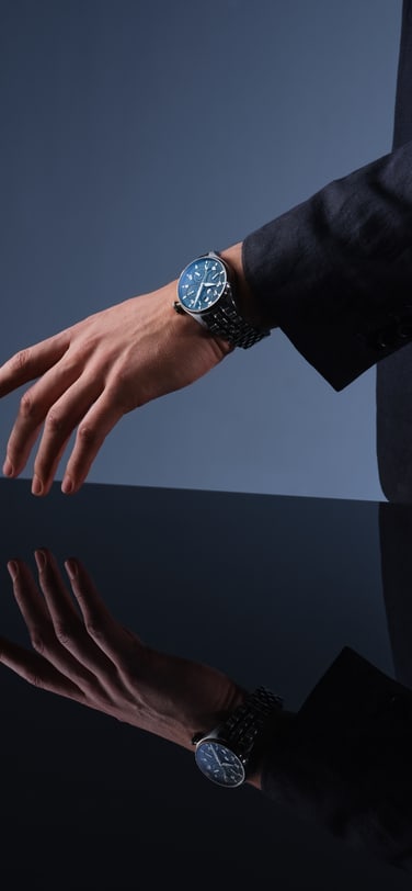 Studio shot of a person in a dark suit with their arm extended on a reflective surface, wearing an IWC watch
