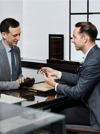 Two elegantly dressed sit around the table holding a watch