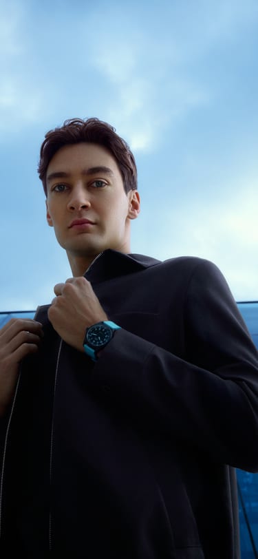 Le pilote de Formule 1, George Russell, en tenue noire, assis devant un bâtiment en verre, portant une montre de sport en céramique noire avec un bracelet bleu