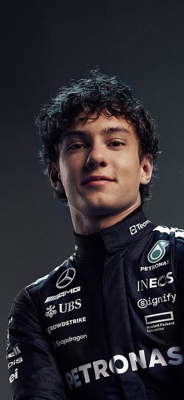 Racer Kimi Antonelli wearing a black racing suit, sitting on a chair and holding a racing helmet