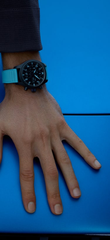 Vue détaillée d’un poignet portant une montre en céramique noire avec un bracelet, des aiguilles et des index bleus contrastés, reposant sur une surface de voiture bleu vif à côté d’un emblème AMG argent.