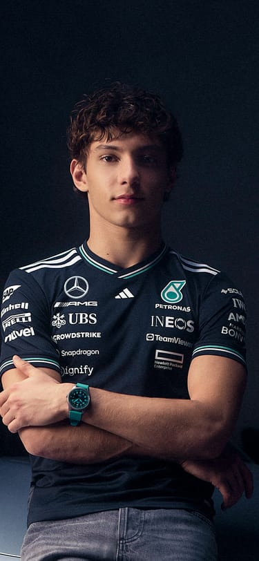 Racer Kimi Antonelli wearing a black racing suit, sitting on a chair and holding a racing helmet