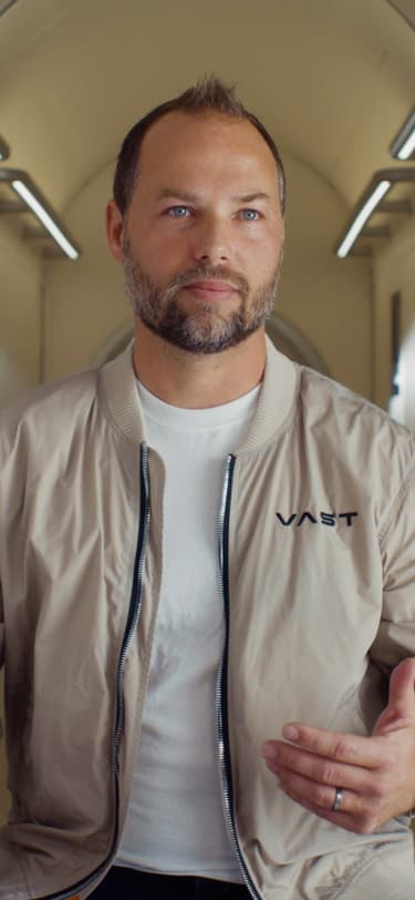 A male Senior Mission Manager wearing a white t-shirt and beige jacket featuring the Vast-logo and flag of the United States.