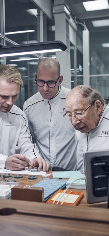 Three watchmakers standing behind a watchmaking table while one explaining the assembly process