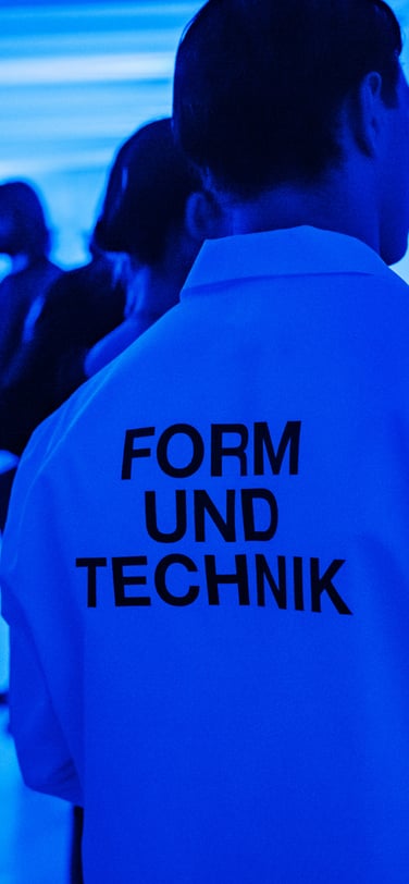 一名男士身穿有「Form und Technik」字樣的T恤，身處打上藍色燈光的展室。