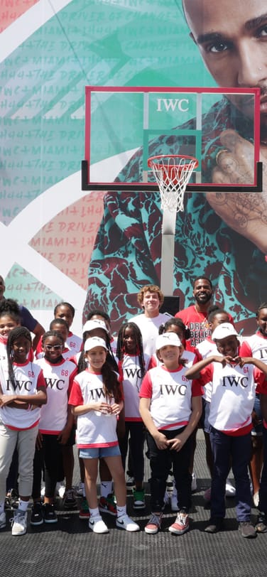 Children and adults in matching IWC shirts pose on an outdoor basketball court in front of a large colourful mural of Lewis Hamilton and hoop.