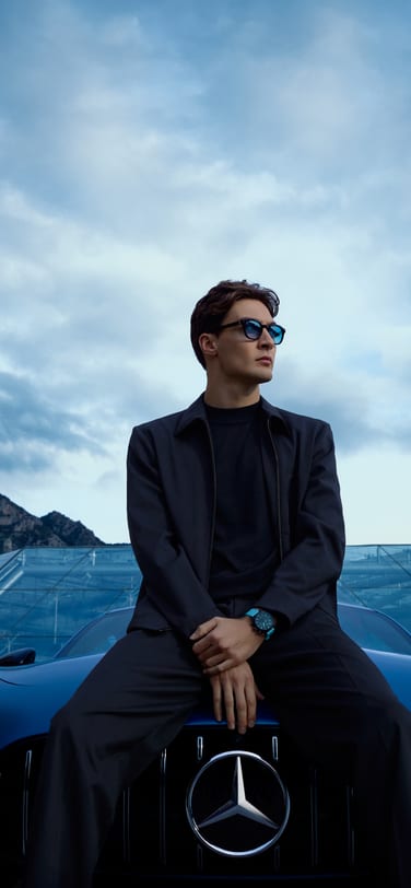 Le pilote de Formule 1 George Russell en tenue foncée avec des lunettes bleues sur une Mercedes-Benz, portant une montre de sport en céramique noire avec un bracelet bleu, avec en toile de fond la ville de Monaco, des montagnes et un bâtiment en verre, sous un ciel nuageux.