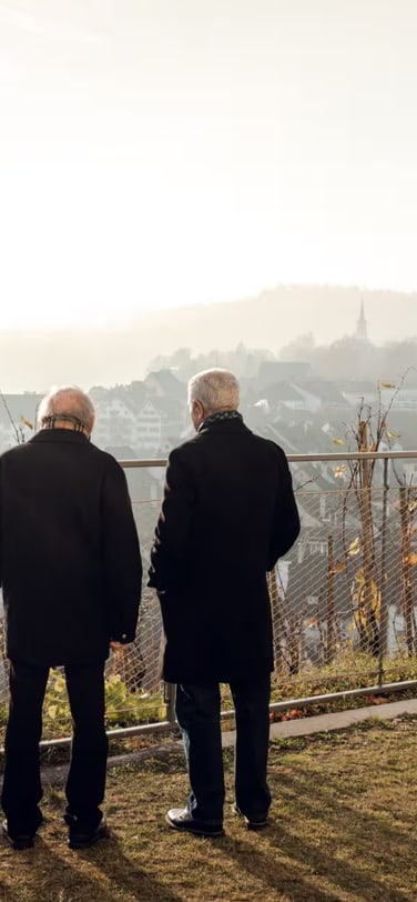 Backshot of watchmakers Kurt Klaus and Amaro Abad dressed in black coats looking over a wire fence