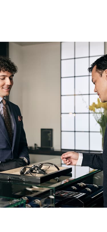 A man shops for luxury watches at a boutique, assisted by a well-dressed salesman behind a glass display case.