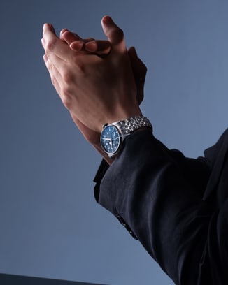 A person in a dark suit with hands clasped, an IWC watch visible on their wrist