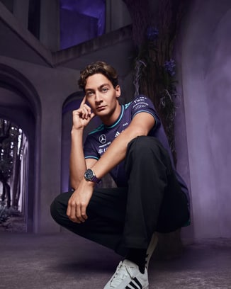 George Russell kneeling wearing wearing Petronas Team shirt, Pilot's Watch Chronograph 41 Mercedes-AMG Petronas Formula One™ Team watch, and Adidas sneakers.